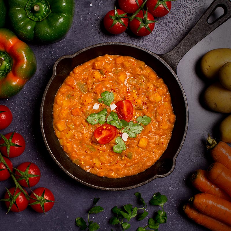 Froyda organic Madras curry in a pan, surrounded by fresh ingredients such as tomatoes, peppers, carrots and potatoes